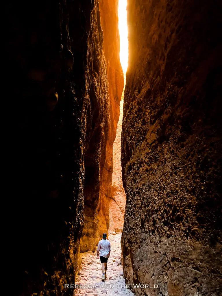 A dramatic, narrow gorge in Purnululu National Park with steep, towering rock walls that create a dark passageway. A lone hiker walks toward a bright opening at the end of the gorge, highlighting the sense of adventure and the park's unique geological formations. This is Echidna Chasm.