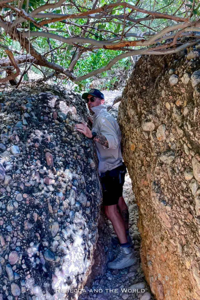 A close-up of a narrow rock crevice in Purnululu National Park, where a man - the author's husband - is carefully squeezing through the tight space between large, weathered rock formations. The rocks are covered with small pebbles and vegetation.
