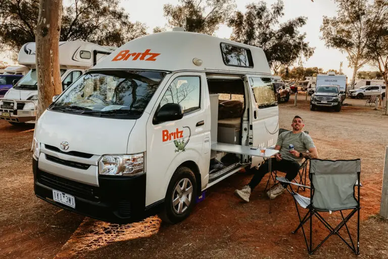 A camper van with the "Britz" logo parked at a campsite with trees around. A man - the author's husband - is seated outside on a camping chair next to a small table, smiling at the camera, enjoying the outdoor camping experience. Renting a campervan in Australia is a great way to see the country.