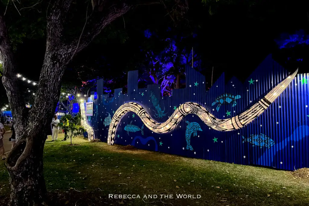 A vibrant night scene featuring a long, illuminated snake artwork displayed on a decorated fence, with people walking around the lit-up area. This artwork was spotted at the Darwin Festival, one of the best reasons to visit Darwin.