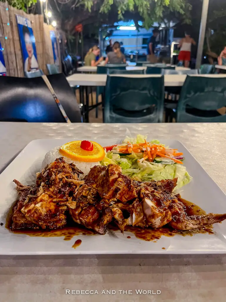 A close-up of a plate featuring a serving of charcoal chicken, white rice and a side salad, set on a table in an outdoor dining area.