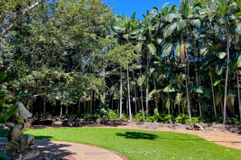 A serene garden scene with tall palm trees and lush greenery, featuring a stone statue of a fish spouting water. Visiting the George Brown Darwin Botanic Gardens is one of the relaxing things to do in Darwin, offering a peaceful retreat within the city’s natural environment - and a break from the city's heat.