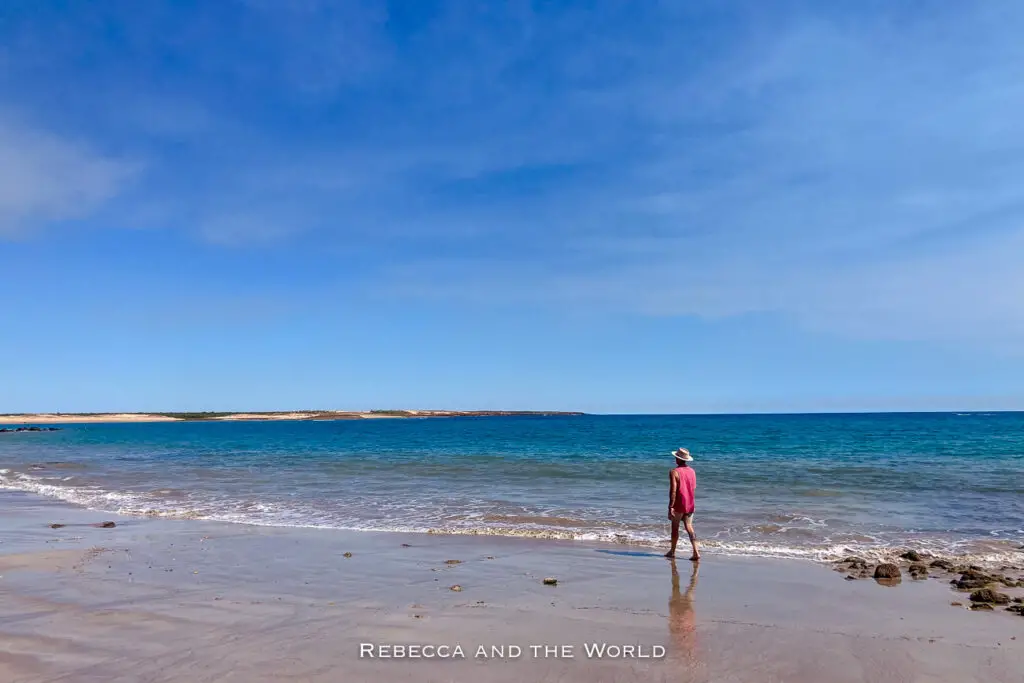 A person - the author's husband - walks along the shoreline of a secluded beach in Dampier Peninsula, wearing a pink tank top and sun hat. Calm waves gently lap at the sandy shore, with the turquoise ocean stretching out under a bright blue sky.