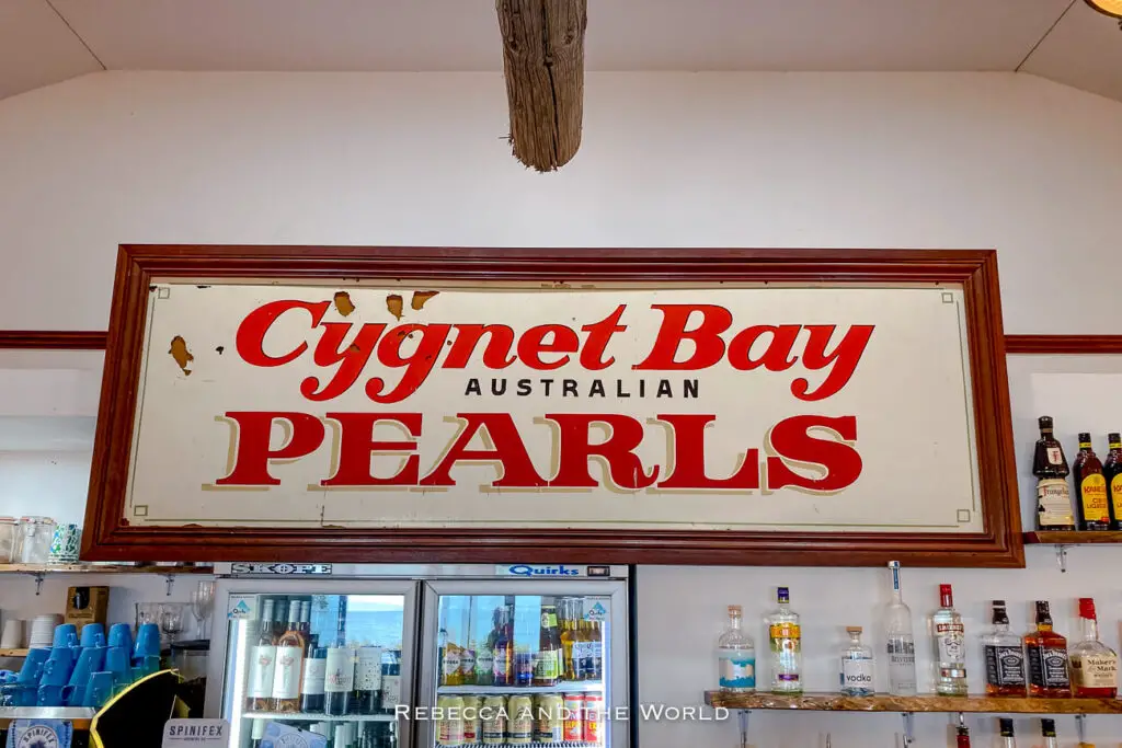 An indoor scene showing the iconic sign for Cygnet Bay Pearls, an Australian pearl farm in the Kimberley region of Western Australia. The vintage-style sign with red lettering is displayed above a bar stocked with various bottles of alcohol, capturing a glimpse of the pearl farm’s visitor centre.