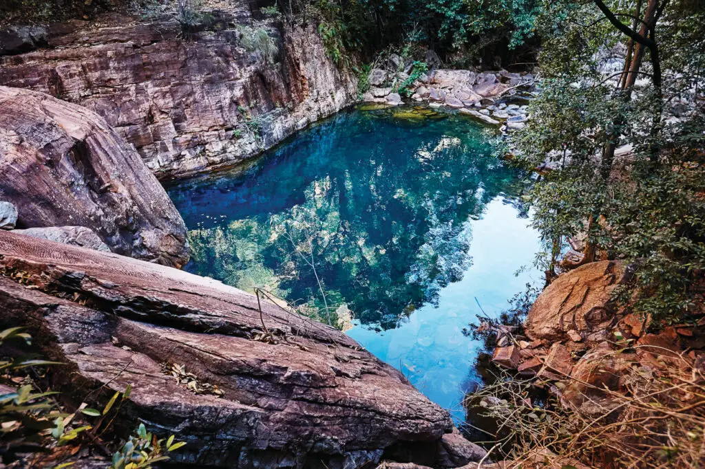 This image depicts a serene, clear waterhole nestled among rocky cliffs and lush vegetation at Emma Gorge. The blue-green water reflects the surrounding landscape, making it a peaceful spot for swimming or relaxation in the Kimberley. Emma Gorge can be found at El Questro, a popular place to visit in The Kimberley region of Western Australia.