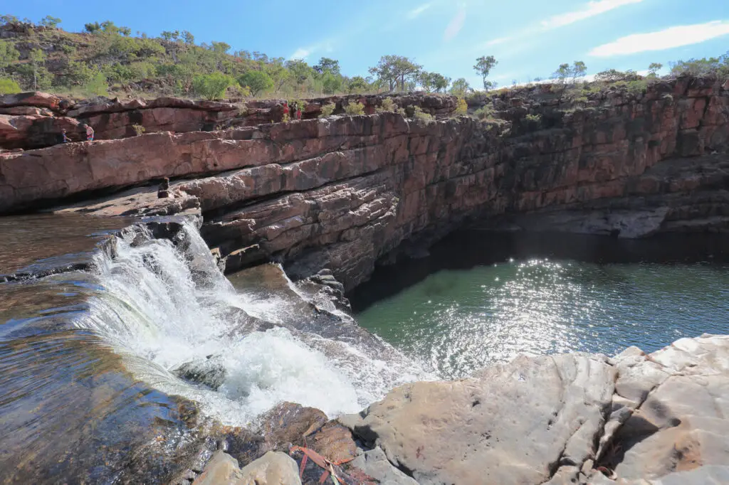 The image shows a waterfall flowing over rocky ledges into a calm, clear pool below at Bell Gorge. The surrounding area is rugged with layers of red rock and sparse greenery, characteristic of the Kimberley landscape. A few people are visible on the rocky outcrop, enjoying the scenic views. Bell Gorge can be found along the famous Gibb River Road in Western Australia's Kimberley region.