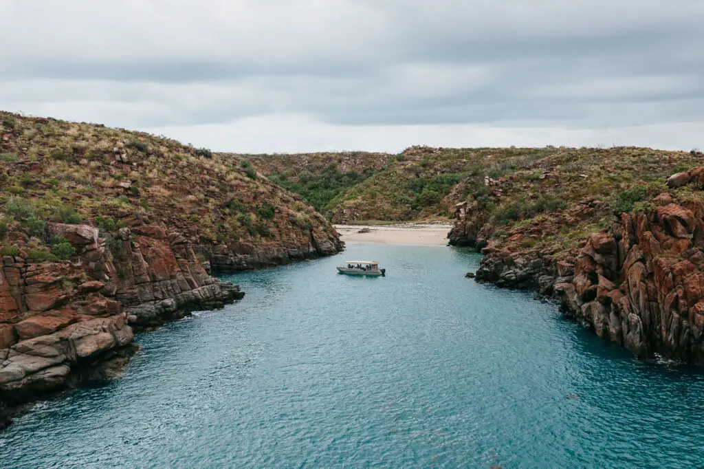 This image captures a small boat navigating through the turquoise waters of a narrow gorge along the Kimberley coastline. The gorge is framed by steep, rocky cliffs covered in rugged vegetation, leading to a secluded sandy beach in the distance.