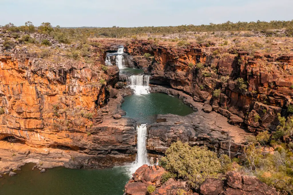 The photo features the multi-tiered waterfalls of Mitchell Falls, surrounded by steep, red rock cliffs and sparse greenery. The waterfalls cascade into a series of pools, creating a dramatic and scenic landscape typical of the remote Kimberley region. Visiting Mitchell Falls is one of the trickier things to do in the Kimberley because it's so remote - but it's worth the effort.