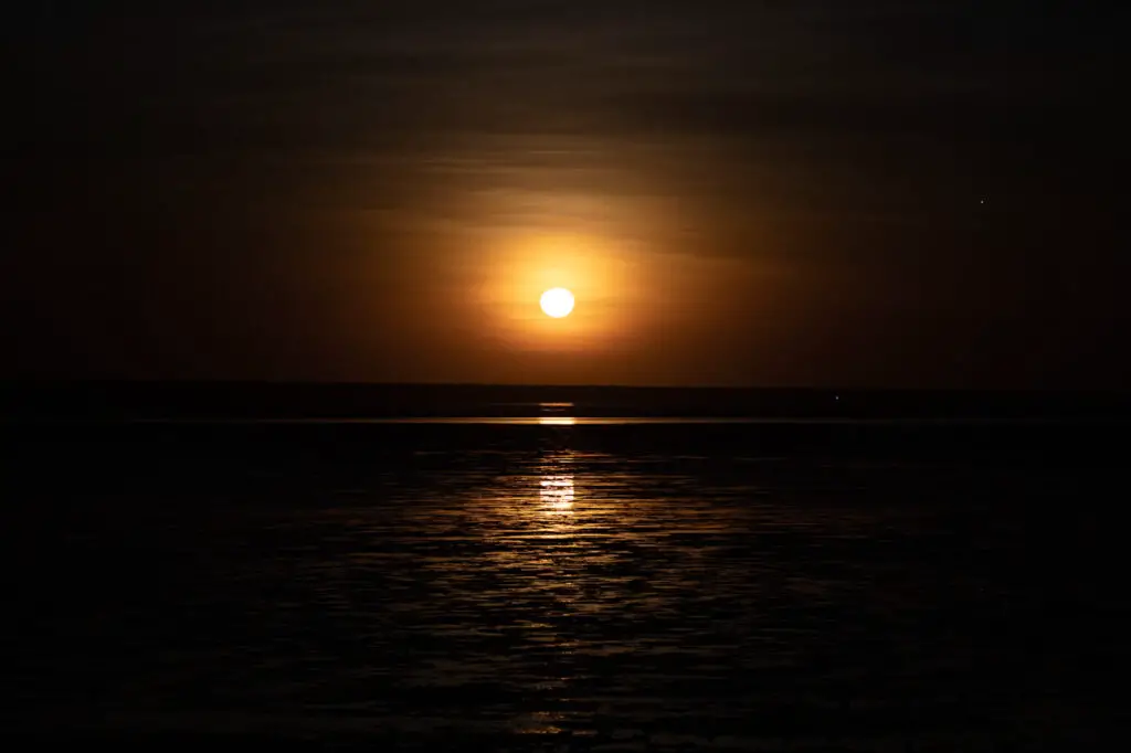 The image captures the natural phenomenon known as the "Staircase to the Moon" in Broome, Western Australia. It shows a full moon rising over the exposed mudflats at low tide, creating the optical illusion of a glowing staircase leading up to the moon. The scene is set against a dark, calm ocean with the moon's reflection shimmering on the water.