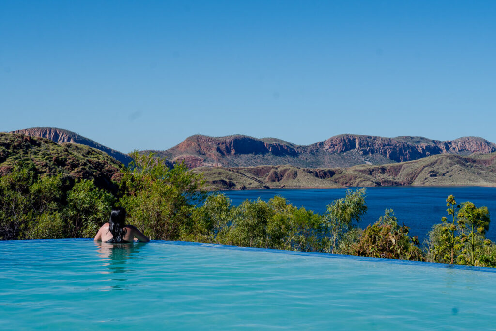 A person - the author of this article - is relaxing in an infinity pool, gazing out over the vast expanse of Lake Argyle in the Kimberley region. The scene captures the serene beauty of the area, with the pool blending seamlessly into the blue waters of the lake, surrounded by rugged hills under a clear sky.
