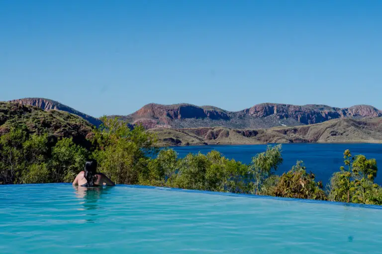A person - the author of this article - is relaxing in an infinity pool, gazing out over the vast expanse of Lake Argyle in the Kimberley region. The scene captures the serene beauty of the area, with the pool blending seamlessly into the blue waters of the lake, surrounded by rugged hills under a clear sky.