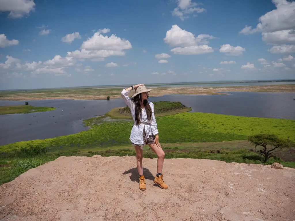 A woman standing on a rocky outcrop, dressed in safari attire, with her hand holding on to her hat as she looks out over a panoramic view of a lush green landscape and a large lake below, typical of the scenic beauty found in Kenya's national parks.