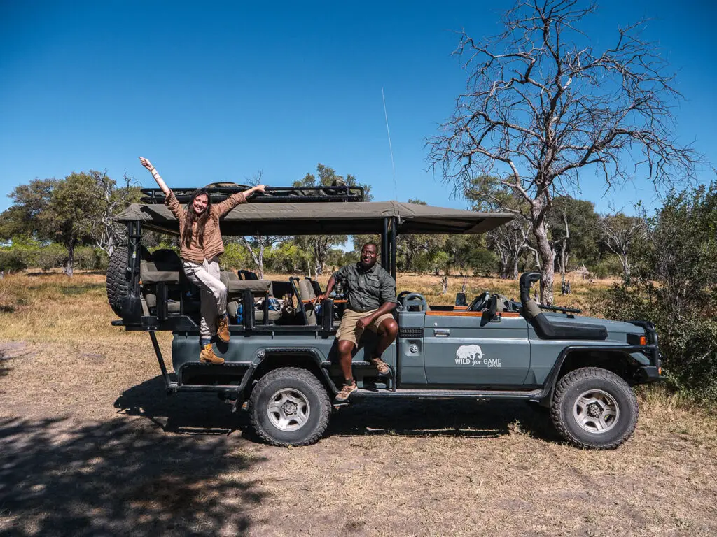Two people sitting on a safari vehicle under a clear blue sky, with one person joyfully raising their arms. The vehicle is parked on a dry, grassy area with sparse trees, highlighting the excitement and adventure of game drives in Kenya's safari parks.