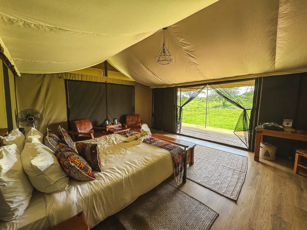 A spacious safari tent with a large bed covered in white linens and patterned pillows. The tent features a view of the surrounding greenery through a wide, open entrance. There are wooden furnishings and a comfortable, cozy ambiance, highlighting the luxury of glamping accommodations available in some Kenyan safari parks.