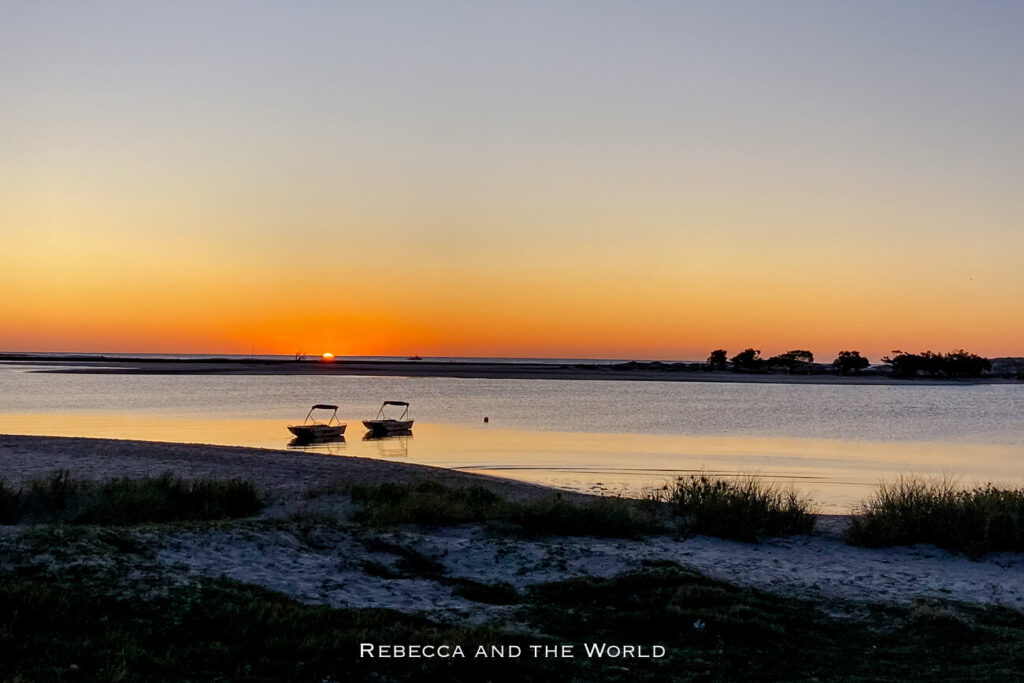 A serene sunset over the water in the town of Kalbarri, Western Australia, with two small boats anchored near the shore. The sun is setting on the horizon, casting a warm orange glow over the calm waters and silhouetting distant trees.