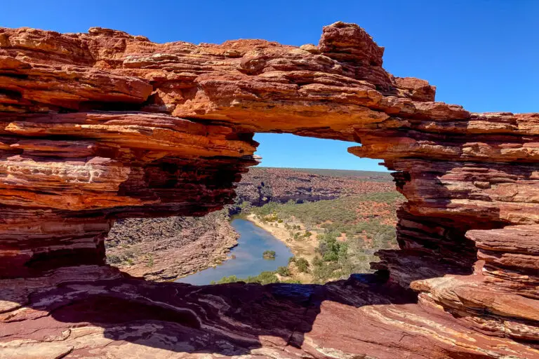 The image features Nature’s Window, an iconic natural rock arch in Kalbarri National Park, Western Australia. The arch, composed of layered red sandstone, frames a view of the meandering Murchison River and the expansive, rugged landscape beyond. The contrasting colours of the red rock, green vegetation, and blue sky create a striking scene, highlighting one of the park’s most famous landmarks.
