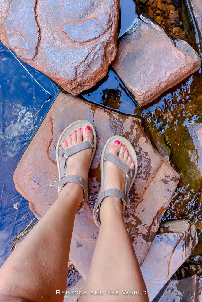 The image shows a pair of feet wearing beige, open-toed sandals with straps, standing on wet red rocks. The toenails are painted red. The rocks are surrounded by shallow, clear water. The view is from a person's perspective (the author of this article's) looking down at their feet. Flip flops or water sandals are a great addition to an Outback Australia packing list.