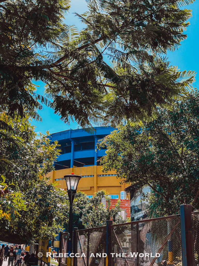 A leafy walkway shaded by tall trees, with sunlight filtering through branches. In the background, a large stadium structure painted blue and yellow - the famous La Bombonera - rises above the greenery. People walk along the path, making it a calm outdoor stop in a Buenos Aires itinerary.