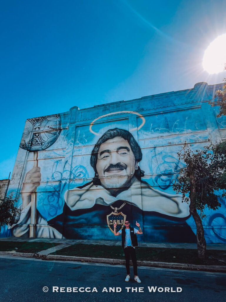 A large mural painted on a blue wall shows Argentine footballer Diego Maradona holding a staff, with a halo-like glow above his head. A person (the author's husband) stands below the mural posing with raised arms. The sun shines brightly in the sky, creating lens flare. Street art like this is a highlight in a 3 days in Buenos Aires trip and a key part of things to do in Buenos Aires.