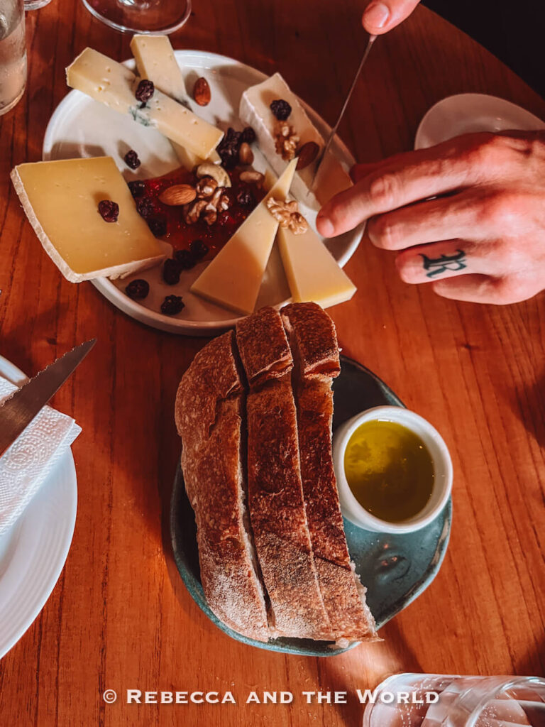 A wooden table with a plate of assorted cheeses, nuts, and dried fruit. In the foreground, slices of crusty bread are served with a small bowl of olive oil. A hand holds a knife over the cheese plate. This is at Pain et Vin, a great wine bar in Buenos Aires.