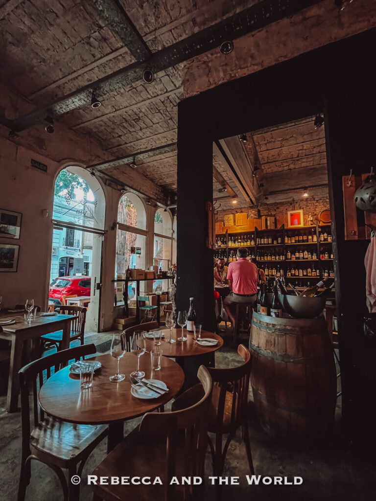 Interior of a cozy restaurant with wooden tables, chairs, and a rustic ceiling. Wine bottles line shelves behind a bar where people are seated. Natural light enters through large arched windows. This is Pain et Vin, a great wine bar in Buenos Aires.