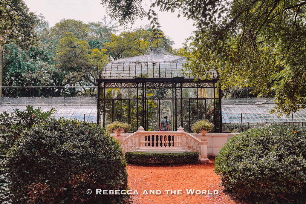 A glass greenhouse surrounded by lush greenery and trees in a landscaped garden. A person walks up steps toward the entrance. The scene is peaceful and well-maintained. It's the Jardin Botanic Carlos Thays, one of the loveliest parks to visit in Buenos Aires.