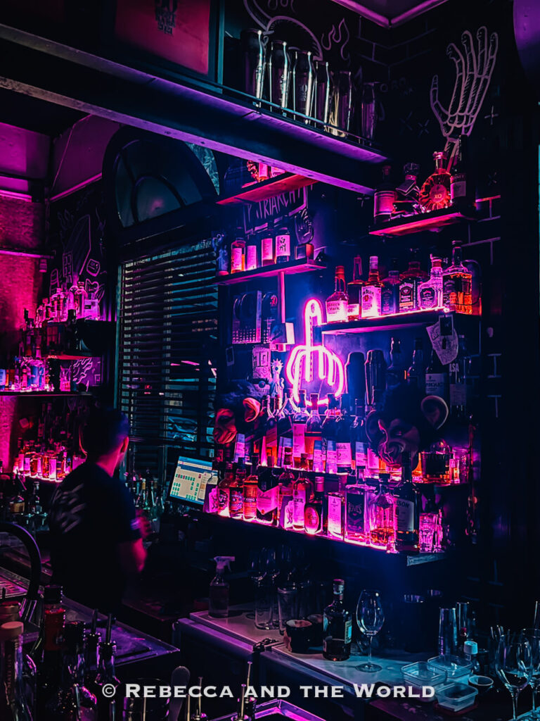 A dimly lit bar interior illuminated by pink and purple neon lights. Shelves behind the bar display various bottles, and a bartender stands at the counter. The vibrant nightlife atmosphere highlights unique things to do in Buenos Aires. Tres Monos is one of Buenos Aires' best bars.