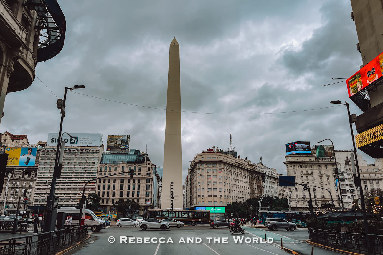 A wide city intersection with traffic, buses, and pedestrians. A tall white obelisk stands in the center under a cloudy sky, surrounded by buildings and billboards. This iconic landmark is a must-see in any 3 days in Buenos Aires itinerary.