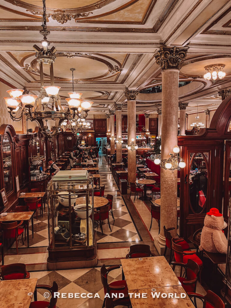 An elegant café interior with high ceilings, ornate chandeliers, marble columns, and checkered floors. Tables and chairs are arranged throughout the space, with people dining. Classic cafés like this - called Confiteria La Ideal - are a staple of things to do in Buenos Aires and often featured in a Buenos Aires itinerary.