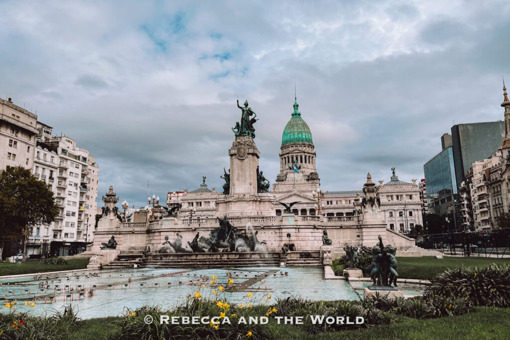 A large historic building with a green dome and ornate statues stands behind a fountain in a public square. Water fills the fountain in the foreground, and the sky is cloudy. This landmark is the Congreso Nacional and it's a key stop in many Buenos Aires itinerary plans and one of the top things to do in Buenos Aires.