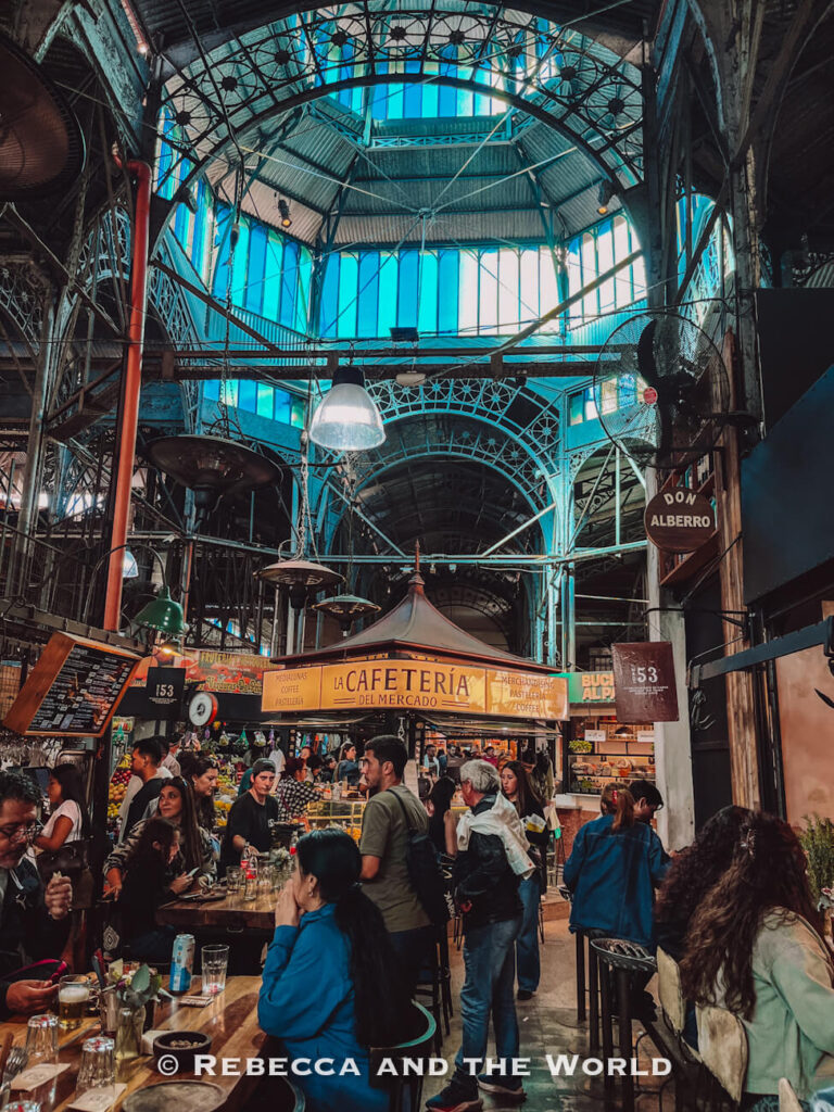 A busy indoor market hall with high ceilings made of metal and glass. People sit at tables and walk between food stalls, including a central café counter. The San Telmo Market is one of the foodie-focused things to do in Buenos Aires during a 3 days in Buenos Aires trip.