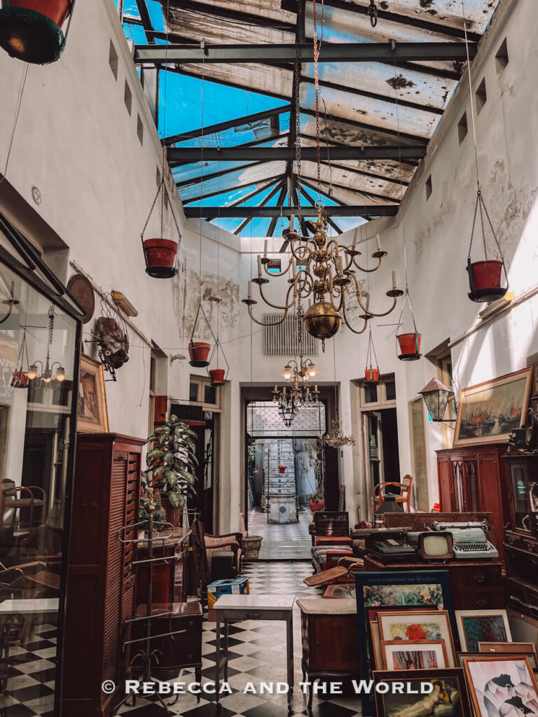 An antique shop interior with vintage furniture, framed artwork, chandeliers, and decorative objects. The space has a glass ceiling and tiled floor, with items arranged closely together. Exploring unique shops like this can be part of a Buenos Aires itinerary.