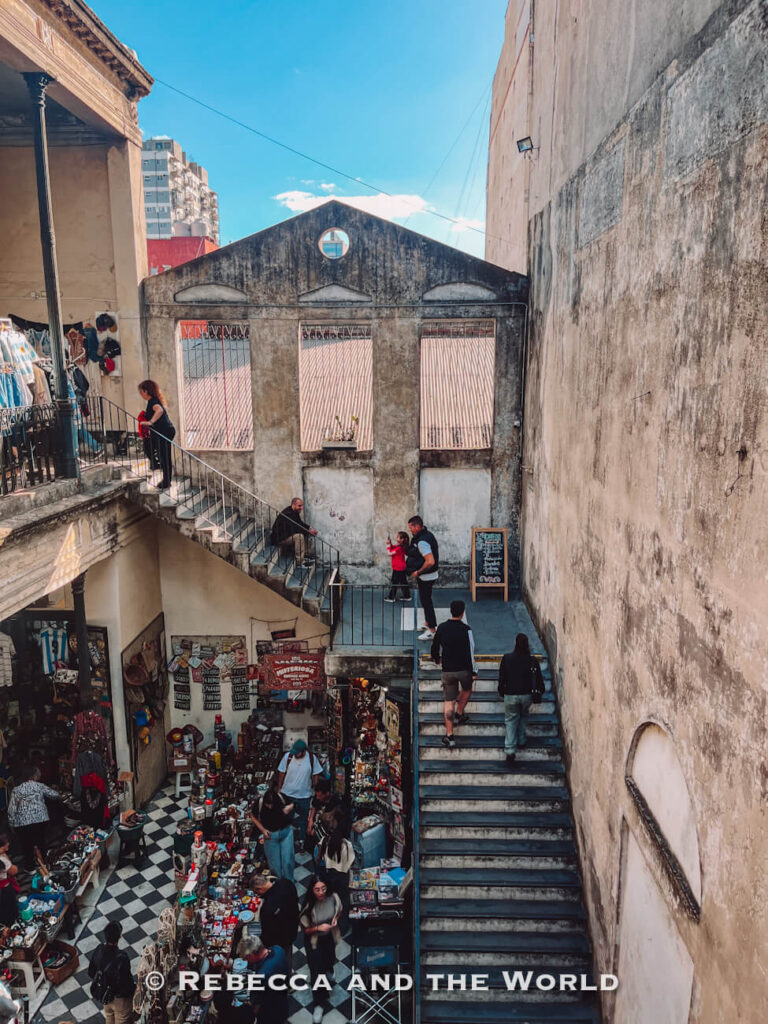 A multi-level indoor market or antique space with staircases and balconies. People walk up and down stairs while browsing stalls filled with various items below.