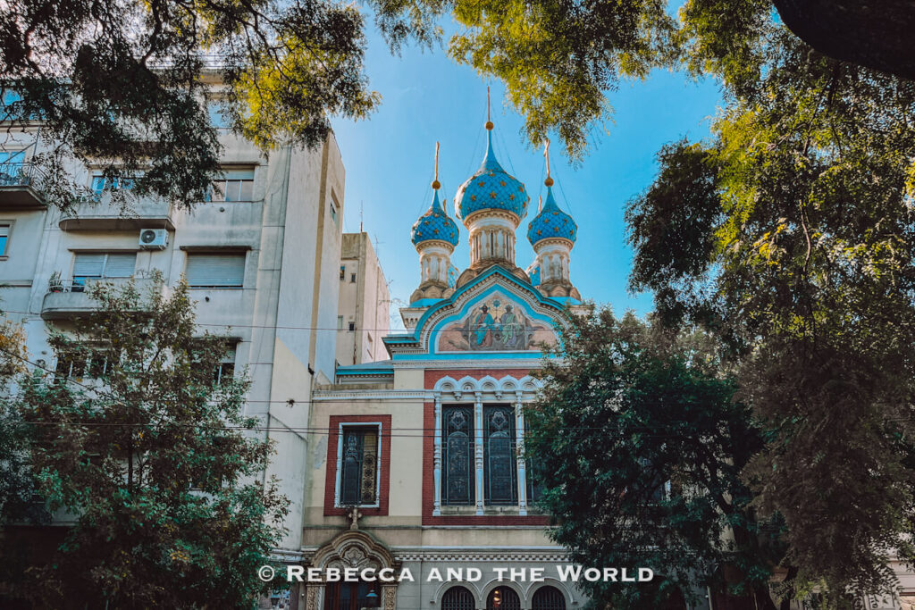 A church with ornate details and distinctive blue onion-shaped domes rises between surrounding buildings and trees. The sky is clear and bright.