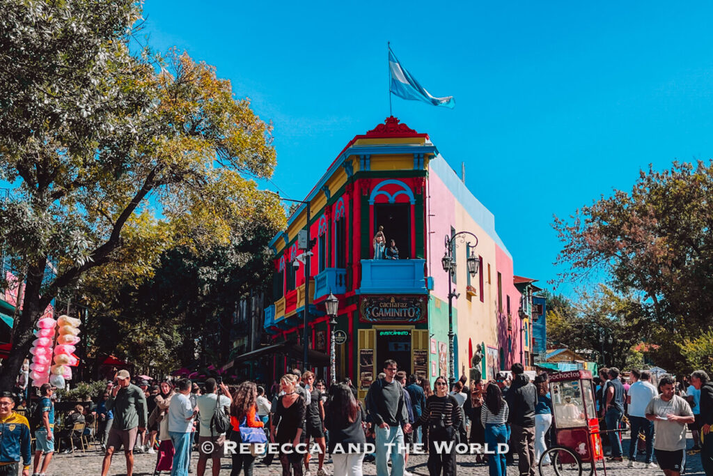 A colorful street corner in La Boca with a bright red, yellow, and blue building. Crowds of people gather around, and vendors sell items nearby under a clear blue sky. This vibrant area is one of the most popular things to do in Buenos Aires.