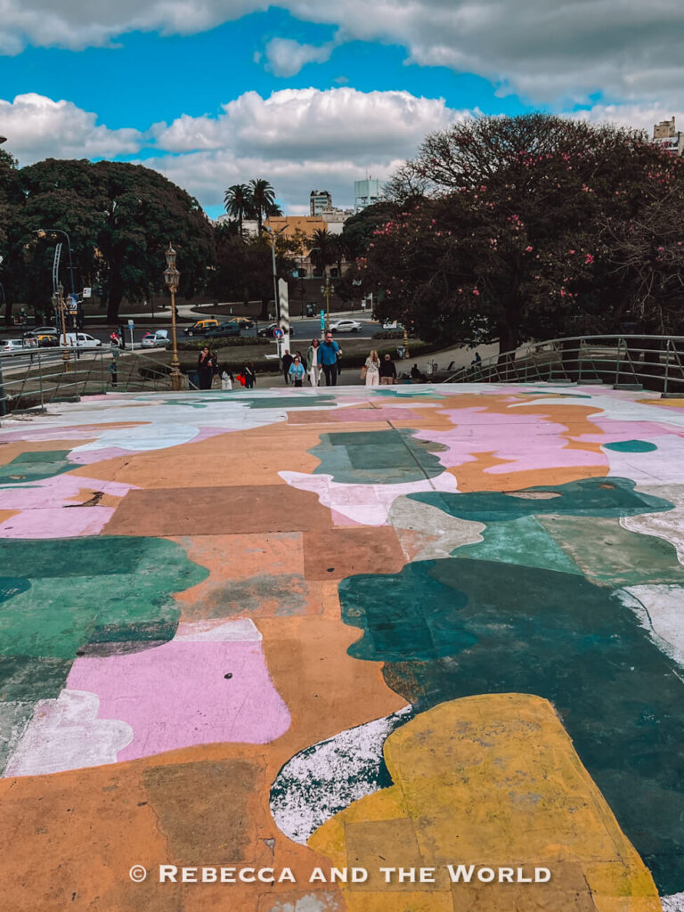 A painted outdoor walkway featuring large abstract shapes in pastel colors. People walk in the background near trees and a street with cars. Public art spaces like this are a creative addition to a Buenos Aires itinerary.