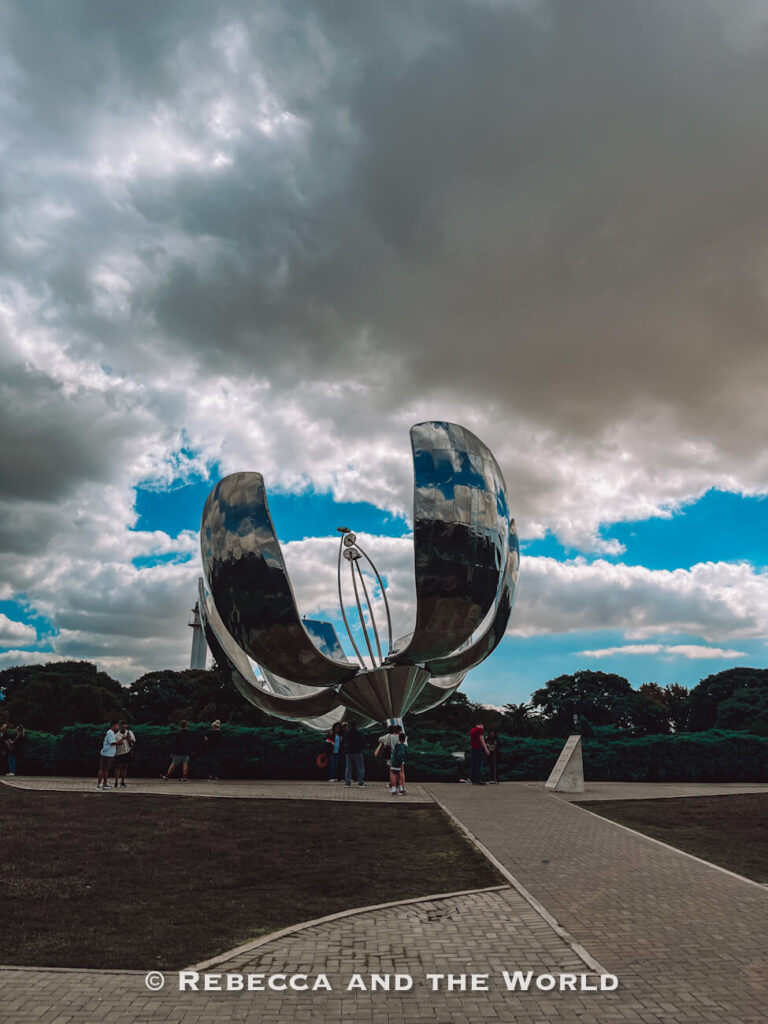 A large metallic flower sculpture stands in a park under a cloudy sky. People gather around the base, and pathways lead up to the structure. Visiting the Floralis Generica can be a great thing to do while spending 3 days in Buenos Aires.