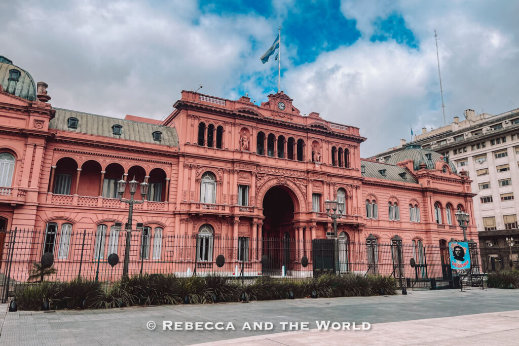 A pink historic building with arches and ornate details stands behind a fence. The Argentine flag flies above the structure under a partly cloudy sky. The Casa Rosada is a must-see when planning things to do in Buenos Aires.