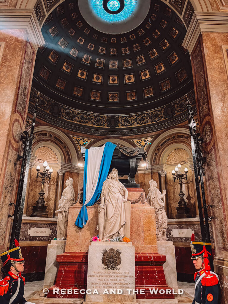 An ornate mausoleum interior with a domed ceiling featuring detailed patterns and a blue skylight. Marble statues surround a central tomb draped with a blue and white cloth, while two guards stand on either side. This is San Martin's tomb inside the Cathedral.