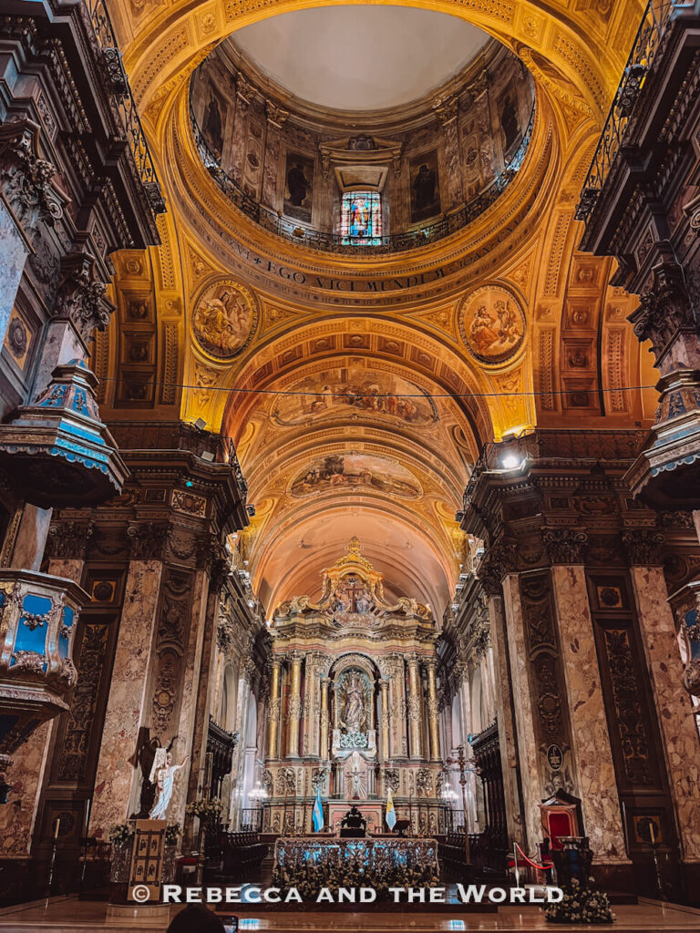 The interior of a grand cathedral with high vaulted ceilings, intricate gold detailing, and religious artwork. An altar sits at the far end beneath a large arch, with soft lighting illuminating the space. Exploring churches like this is a common stop in a Buenos Aires itinerary.