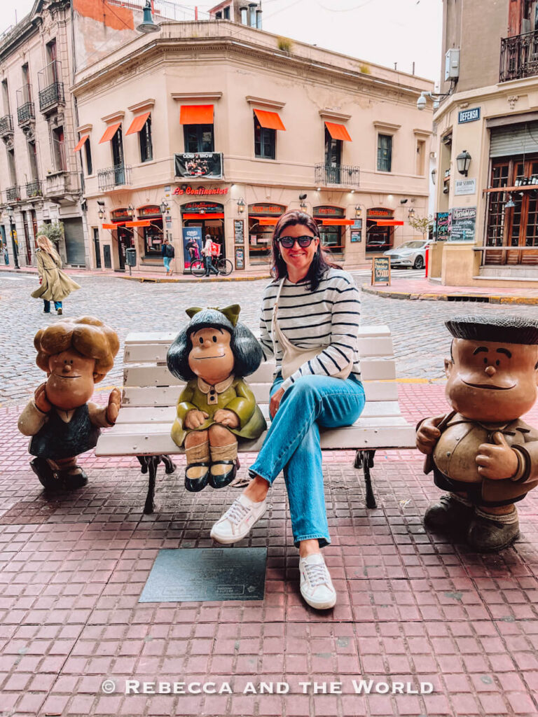 A woman sits on a bench beside cartoon-style statues of children on a cobblestone street corner. Behind her, a café with orange awnings faces the intersection. This playful photo spot with beloved cartoon character Mafalda is one of the fun things to do in Buenos Aires during a 3 days in Buenos Aires trip.