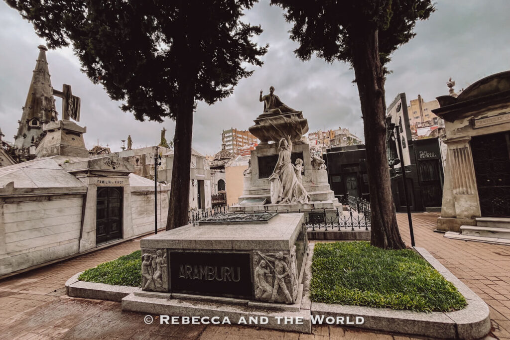 A cemetery scene with ornate stone mausoleums, statues, and monuments arranged along paved walkways. Trees line the area, and the sky is overcast. Visiting the Recoleta Cemetery is one of the more unique things to do in Buenos Aires.