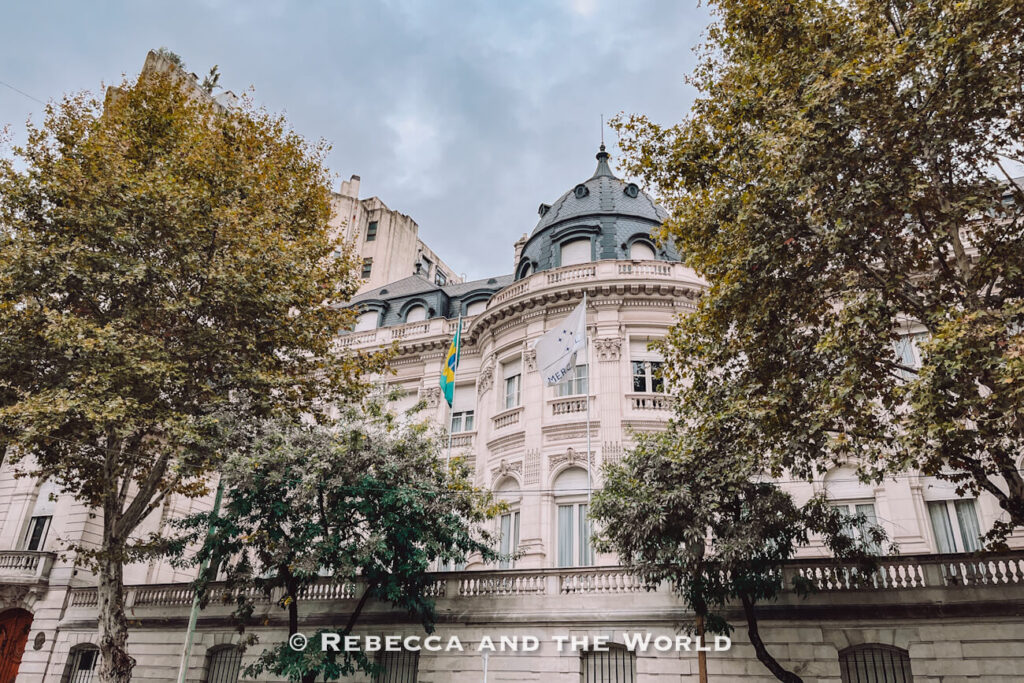 A large historic building with a rounded dome and decorative facade stands behind trees. Flags hang from the front, and the sky is cloudy. Architectural landmarks like this are often featured in a 3 days in Buenos Aires itinerary.