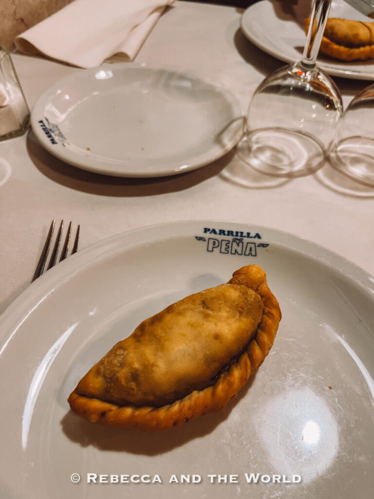 A single empanada sits on a white plate on a restaurant table, with glasses and cutlery nearby. The setting suggests a casual dining experience. Trying traditional foods like empanadas is a must-do when visiting Buenos Aires.