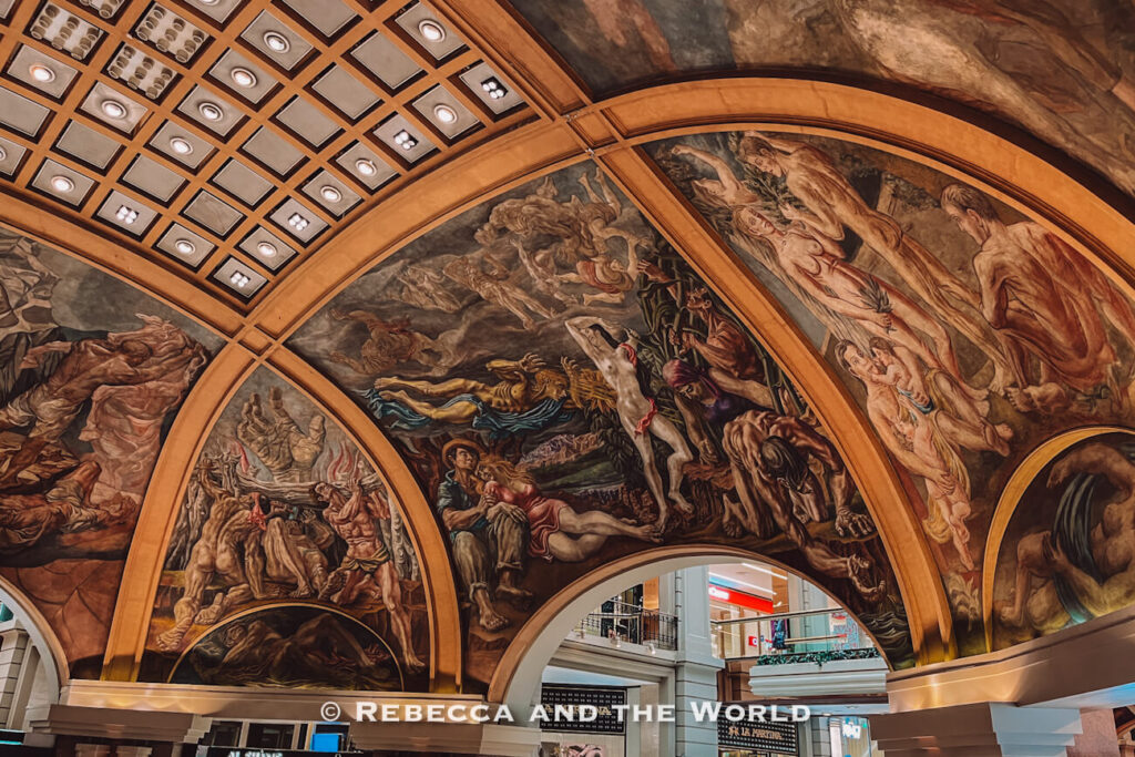 A ceiling covered in large, detailed murals depicting human figures and dramatic scenes, framed by arched architectural elements. Decorative lighting panels are visible alongside the artwork. This is the Galerias Pacifico shopping centre.