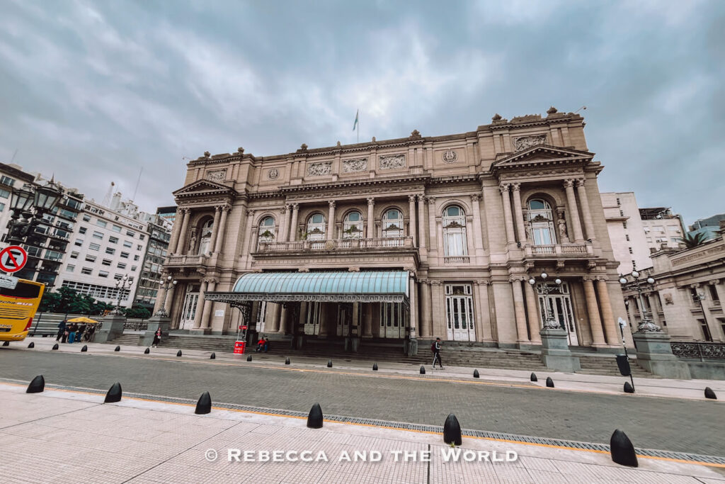 The exterior of a grand historic theater with columns, arches, and ornate detailing. The building faces a wide street with a few pedestrians nearby under a cloudy sky. I always recommend visiting the Teatro Colon when on a 3 days in Buenos Aires itinerary.