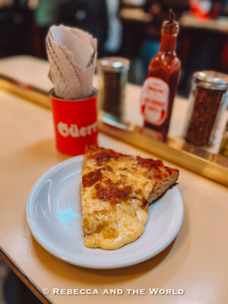 A slice of cheese pizza sits on a small white plate on a café counter. Behind it are condiments, including a bottle of sauce and spice shakers, along with a container holding folded napkins.