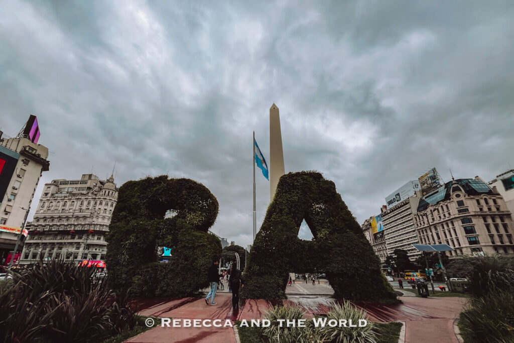 Large sculpted letters “BA” made from greenery stand in a public square. Behind them rises a tall white obelisk, with city buildings and a cloudy sky in the background.