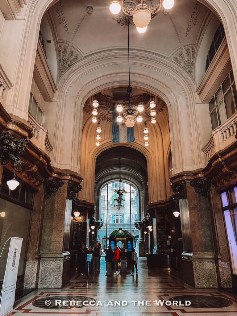 An elegant interior hallway with high arched ceilings, hanging chandeliers, and polished floors. Several people stand near the entrance at the far end, and an Argentine flag hangs from the ceiling. The Palacio Barolo is one of the more unique things to do in Buenos Aires.