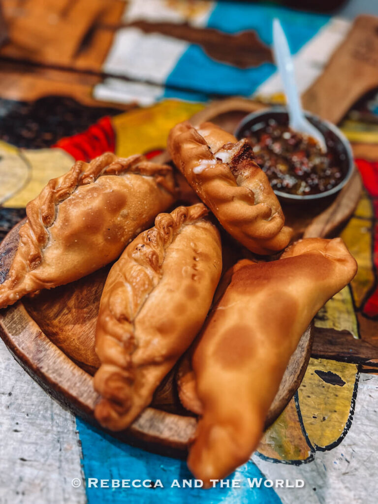 A wooden plate holds several golden-brown empanadas arranged in a circle. A small bowl of dipping sauce sits beside them on a colorful tabletop. El Gauchito is one of the best places to eat in San Telmo.